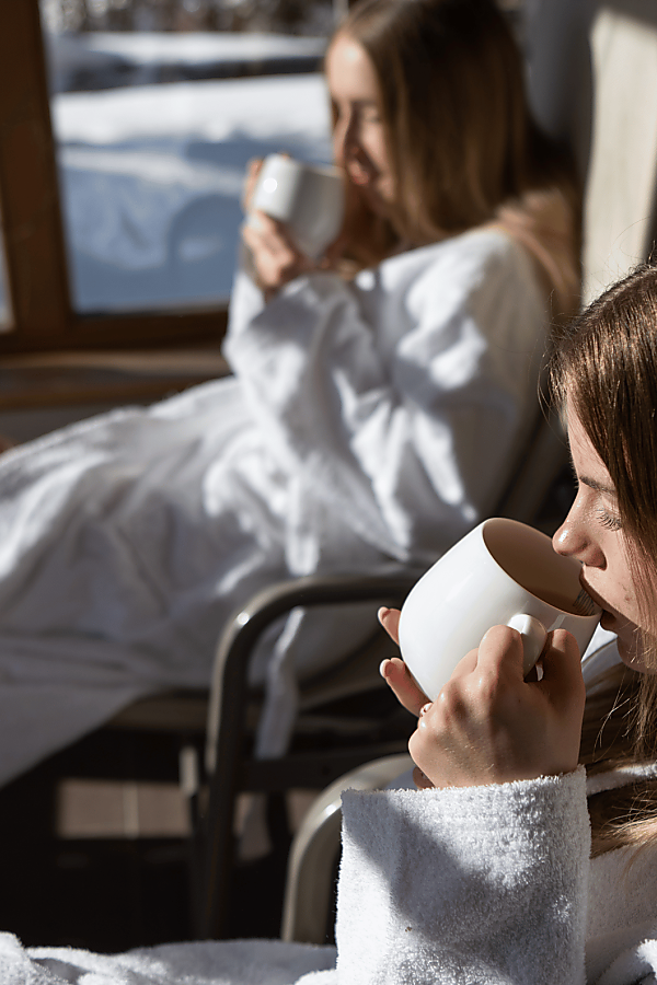 Winterruhe im Bademantel – Retreat-Auszeit in der Almlust Zwei junge Frauen entspannen im Bademantel mit einer heißen Tasse Tee im sonnigen Ruhebereich der Almlust