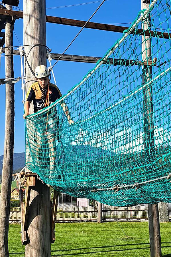 Kinder im Hochseilgarten Flachau – sicheres Abenteuer in luftiger Höhe Kind mit Helm im Kinderhochseilgarten Flachau balanciert über grüne Netzbrücke zwischen Holztürmen bei blauem Himmel